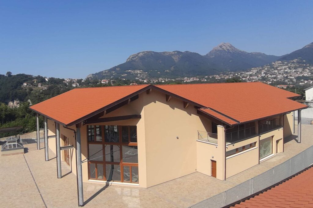 Struttura esterna di colore ocra con tettoie rosse a spiovente che ospiterà la nuova Cantina Marisa Cuomo ad Agerola che servirà vino in Campania. Sullo sfondo s'intravede una dorsale montuosa.  